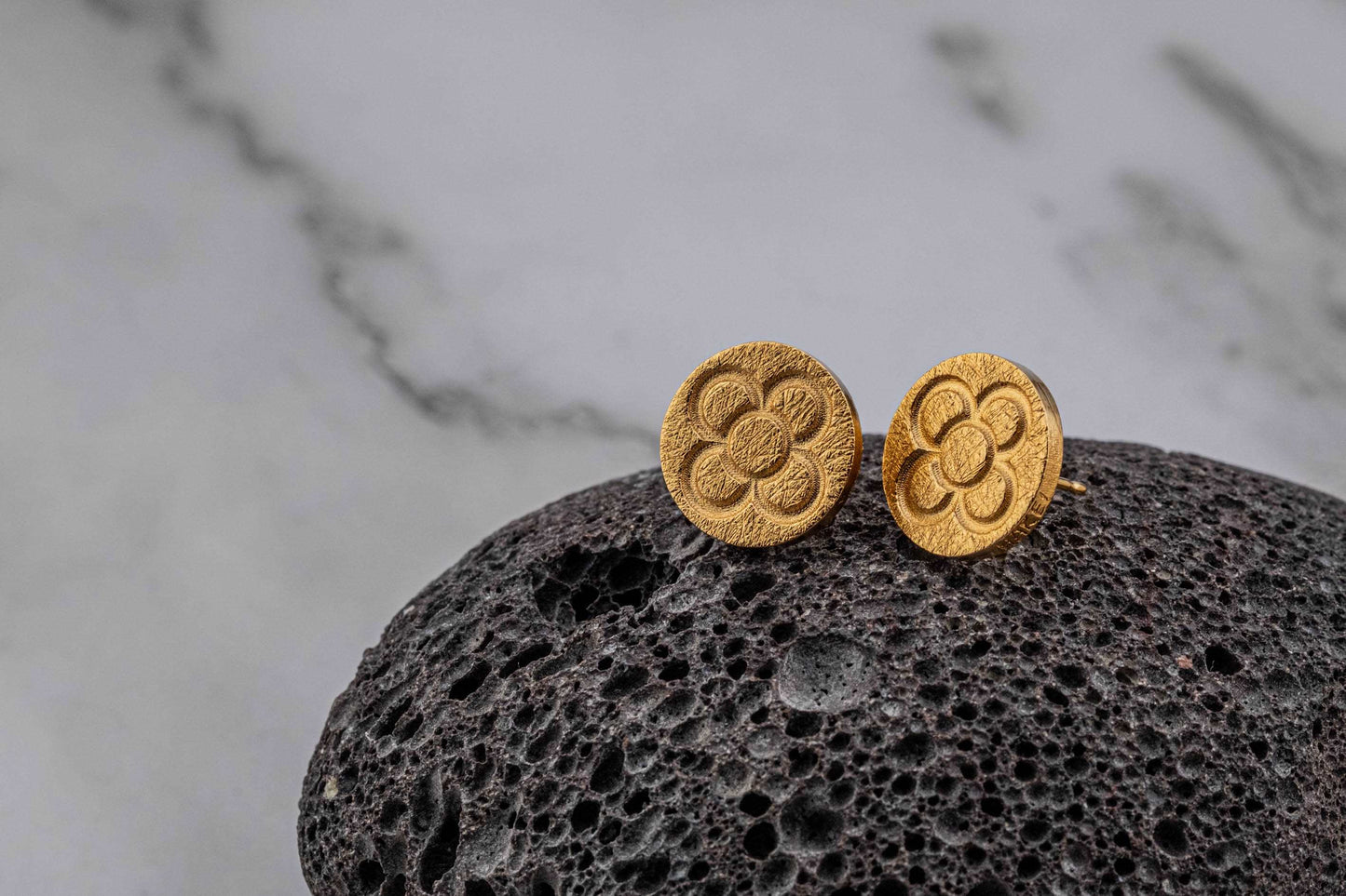 Gold round stud earrings with raised flower pattern on dark textured stone
