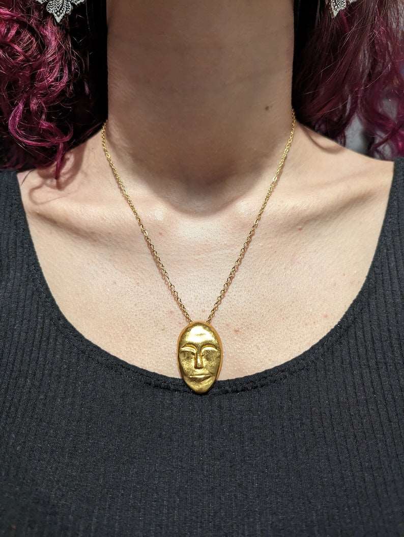Gold face-mask pendant necklace on a collarbone with a fine chain link