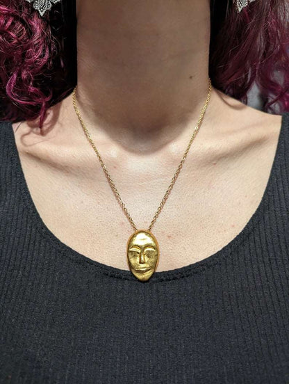 Gold face-mask pendant necklace on a collarbone with a fine chain link